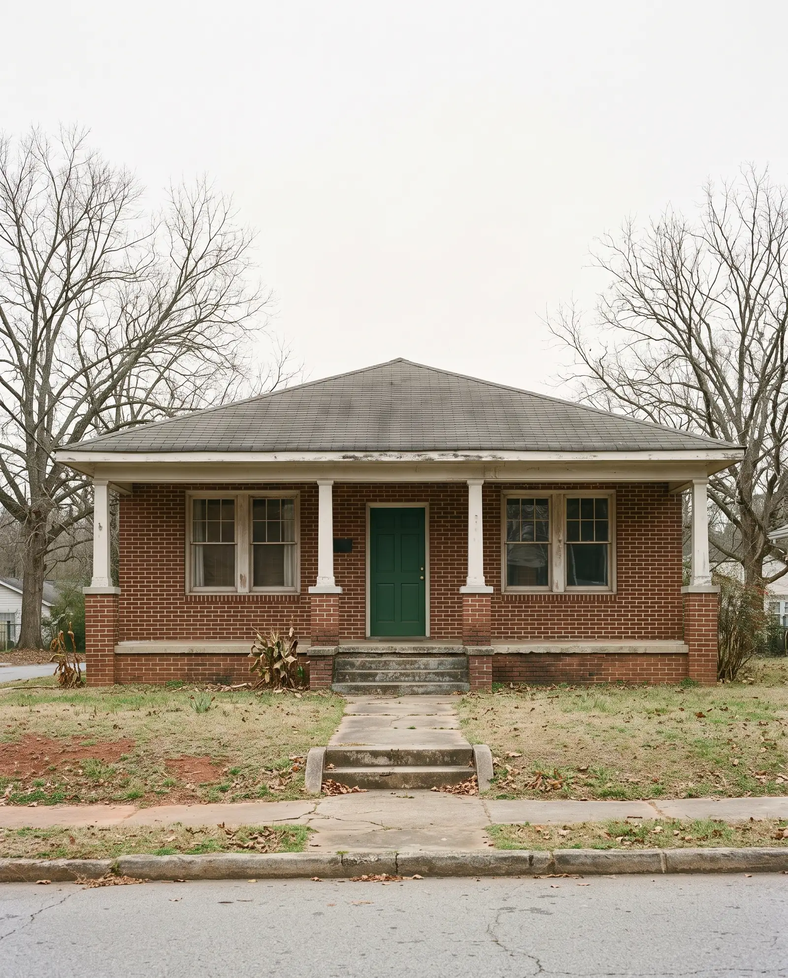 A 1940s brick bungalow with a deep front porch and a green front door, seen across a quiet street in November.